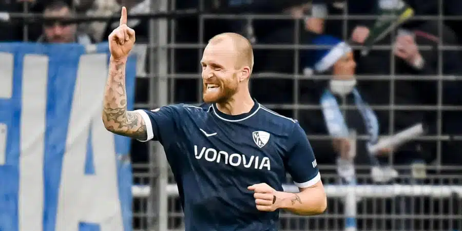 Soccer player in a dark blue kit raises one finger in celebration on the pitch, with fans behind a fence in the stands.