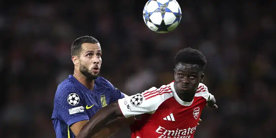 Two players contest a header in a soccer match, one in blue and yellow and the other in red and white, as the ball sails above them.