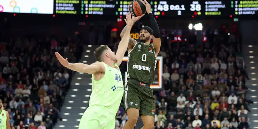 Basketball player in dark green jersey leaps for a shot while a defender in neon green reaches to block in a crowded arena.