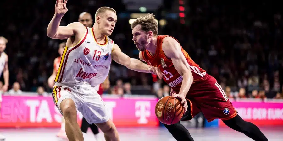 Basketball player in white defends as a red-jersey opponent dribbles the ball toward the basket amid a crowded arena.