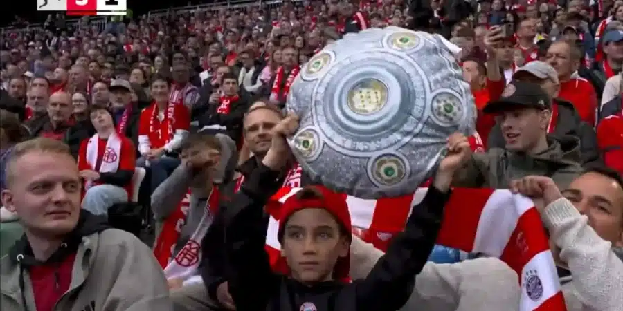 Young boy wearing a red headband raises a large round silver trophy amid a sea of red-and-white-clad fans in the stadium.