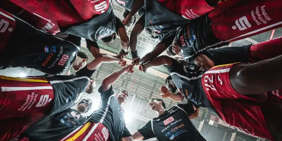 Team of athletes in red and black uniforms form a circle, hands joined in the center to show teamwork and unity in an indoor arena.