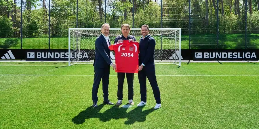 Three men in suits stand on a grass soccer field, presenting a red jersey with the number 2034 in front of a goal and Bundesliga banners in the background.