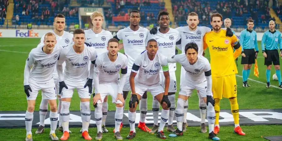 Soccer team in white jerseys posing for a group photo on the field, with the yellow-clad goalkeeper on the right and stadium seats in the background.