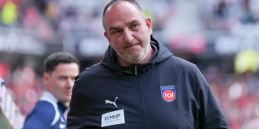 Head-and-shoulders of a middle-aged man in a dark blue sports jacket with a club badge, standing on a football pitch.