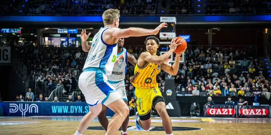 Basketball player in yellow drives with the ball while a defender in white blocks in a crowded indoor court.