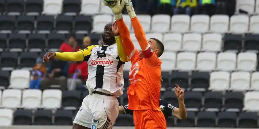 Soccer players jump for a high ball; the goalkeeper in orange reaches up to punch the ball while a white-jersey defender challenges nearby in a stadium.