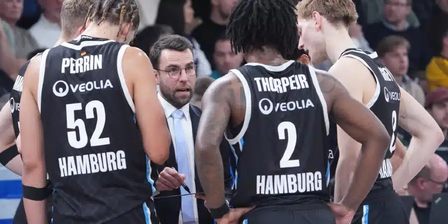 Basketball coach in a suit speaks to Hamburg team players during a timeout, players in black jerseys with Veolia logos and numbers 52 and 2.