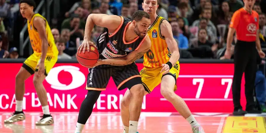 Basketball player in a black-orange jersey dribbles the ball while a defender in a yellow-striped jersey guards him on a crowded court with bright advertising boards behind.