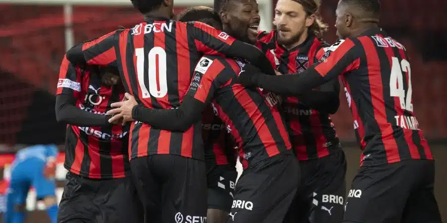 Soccer players in red and black striped jerseys celebrate with a group hug on the field.