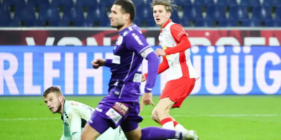 Soccer players chase the ball during a match; a purple-uniform player leads as a red-and-white player pursues, while a light-green kit is on the ground nearby.
