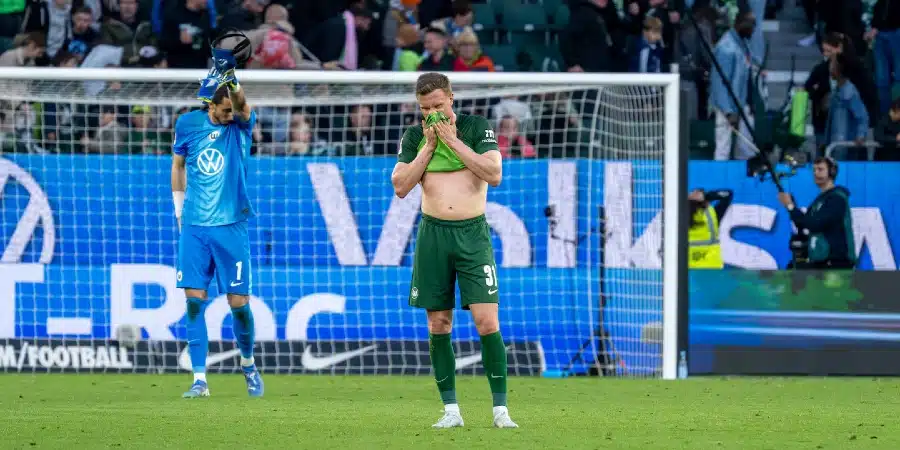 Soccer scene: a green-uniform player pulls his shirt over his head near the goal, while a blue-uniform goalkeeper stands nearby as spectators watch from the stands.