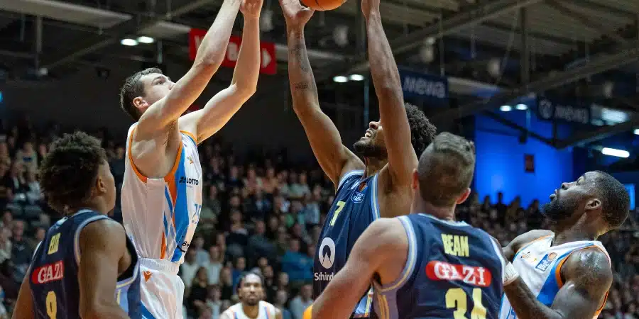 Basketball players jump and reach for the ball near the basket as defenders contest a shot, with a crowded arena in the background.