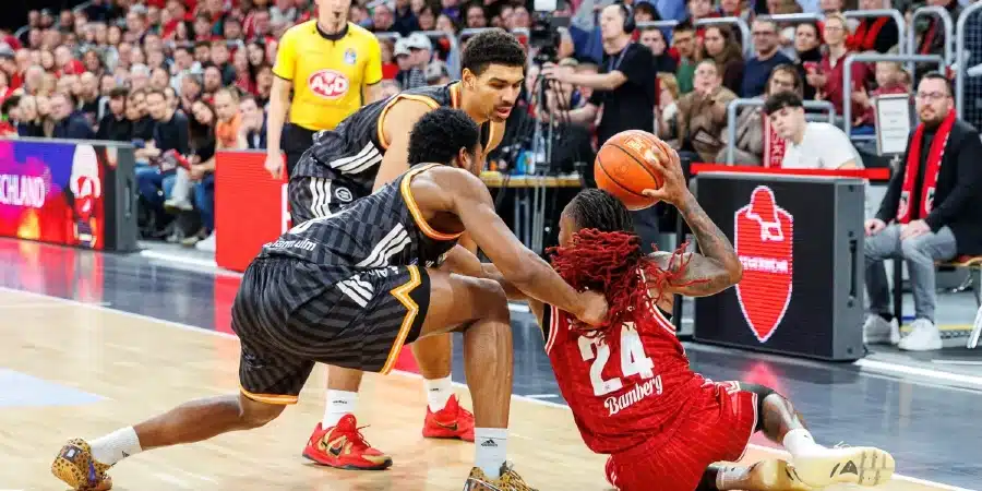 Basketball scene with a player in red on the floor reaching for the ball while two defenders in black close in; crowded stands in background.