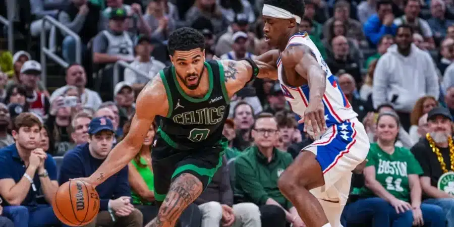 Two basketball players compete for the ball near the baseline with a crowded arena in the background, one wearing a Celtics green uniform dribbling as the defender challenges him.