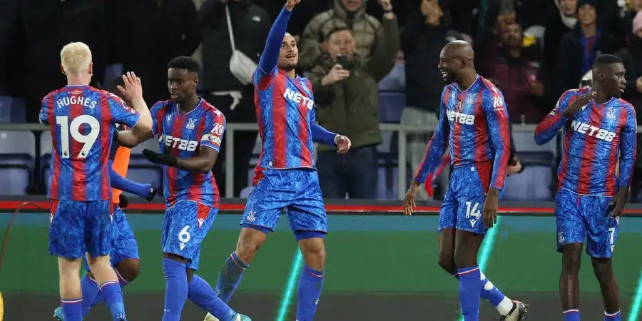 Five football players in blue-and-red striped kits celebrate on the pitch, high-fiving as fans cheer in the stands behind them.