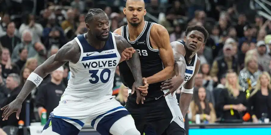 Basketball players fighting for the ball during a fast-paced game, with a Wolves player in white and blue challenging a defender in black gear as fans watch intently in the background.