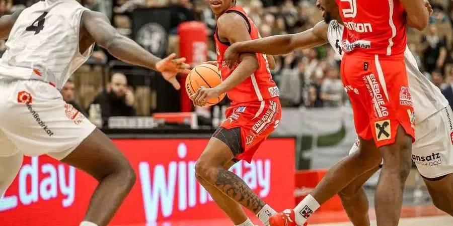 Basketball player in red drives with the ball while defenders in white reach in.