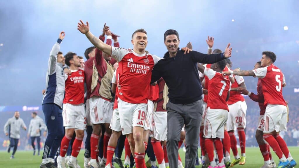 Arsenal players celebrate with manager on the pitch after a win, arms raised in jubilation.