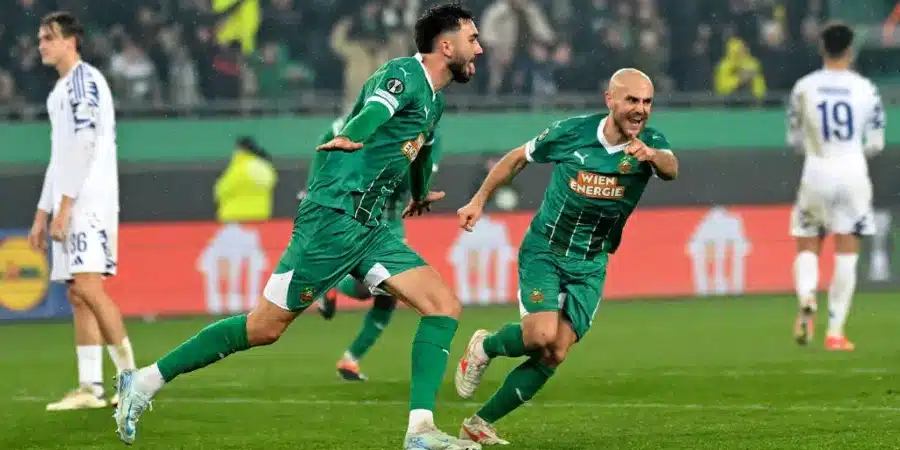 Two players in green jerseys sprint and celebrate a goal on a soccer pitch, while a white-clad opponent watches in the background.