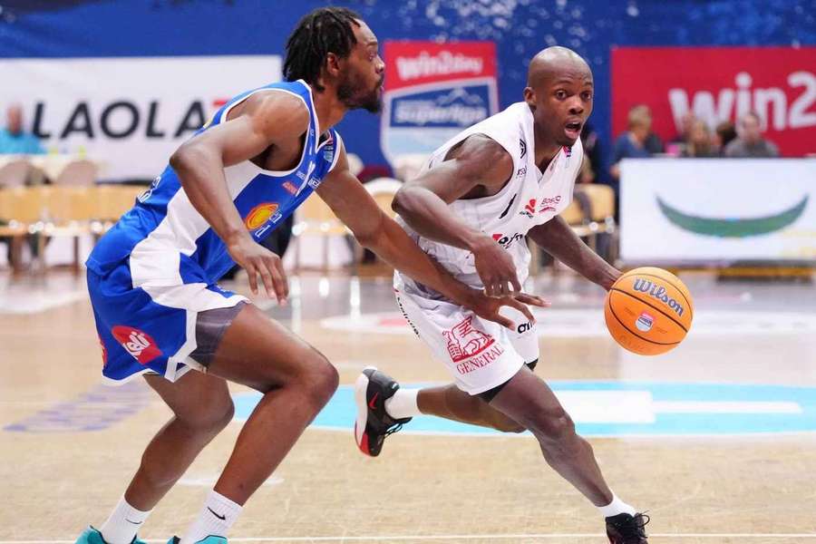 Two basketball players chase a dribbling ball on an indoor court, one in white ahead of a defender in blue.