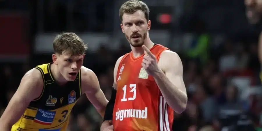 Two basketball players compete on court during a game; foreground player wears red jersey with number 13, pointing his finger while the yellow‑black jersey player stands beside him and blocks the view into the arena.