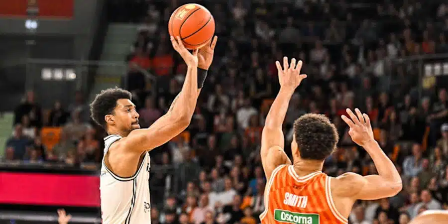 Basketball player in white jersey shoots while defender in orange jerseys raises hands to block in a crowded arena.