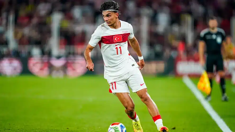 Turkish national team footballer wearing number 11 in a white kit with red stripe dribbles the ball on a green pitch during a match.