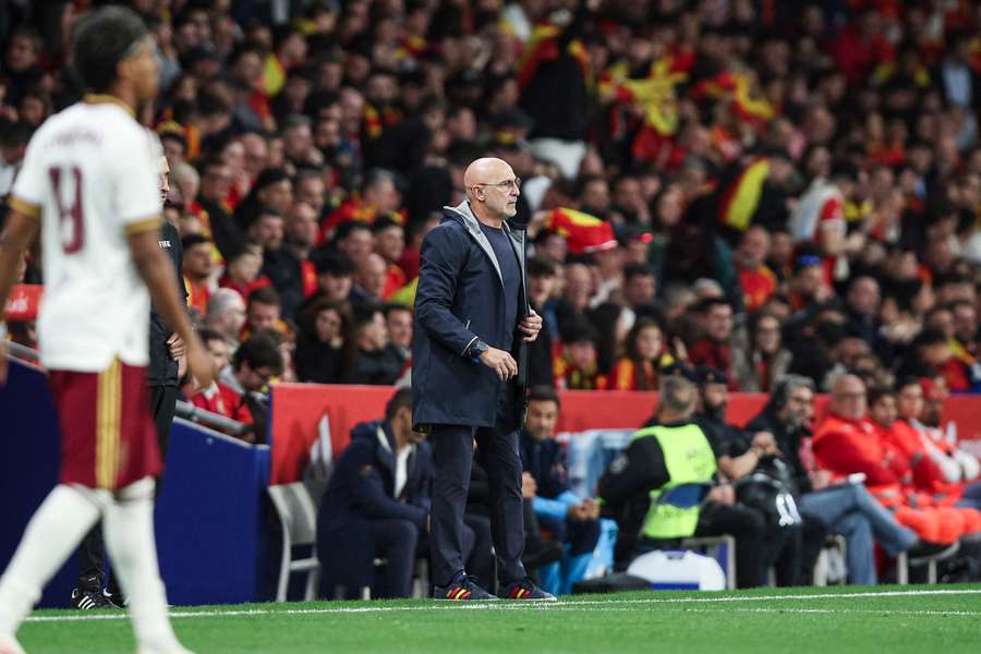 Bald man in a dark coat stands on the soccer sideline during a match, with red-and-yellow-clad fans in the stands behind him.