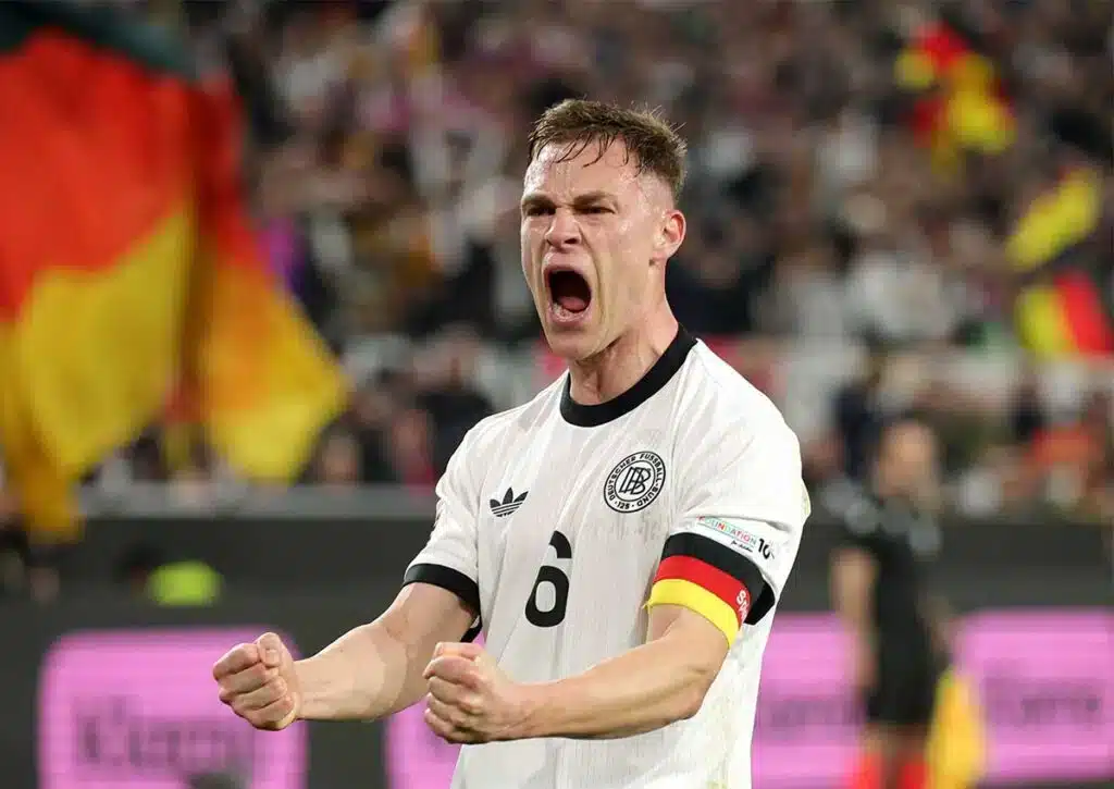 Soccer player in a white Germany jersey celebrates with clenched fists and mouth open on the pitch, armband visible on arm.