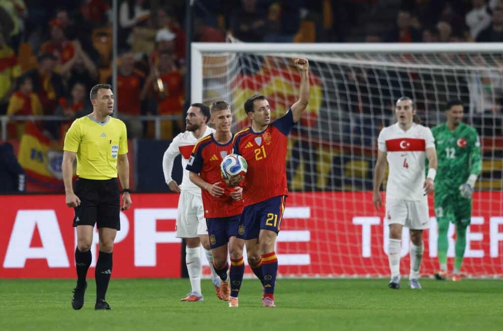 Soccer player in red and blue uniform raises his fist in celebration near the goal, holding the ball as officials and teammates look on.